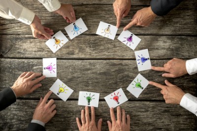 Teamwork and brainstorming concept with businessmen seated around a table each pointing to cards with colorful sketches of light bulbs conceptual of bright ideas and solutions arranged in a circle.